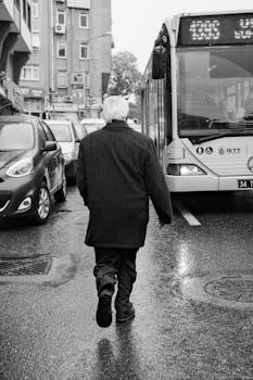 Black and white photo of an elderly man walking near a bus on a wet city street.