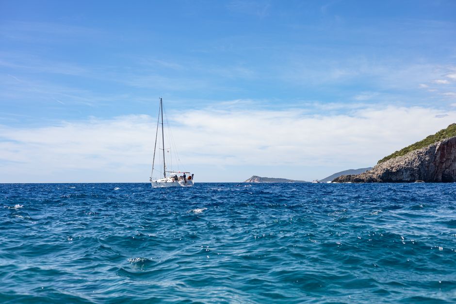 A serene view of a yacht sailing on the open ocean with a rocky coast in the distance under a clear blue sky.
