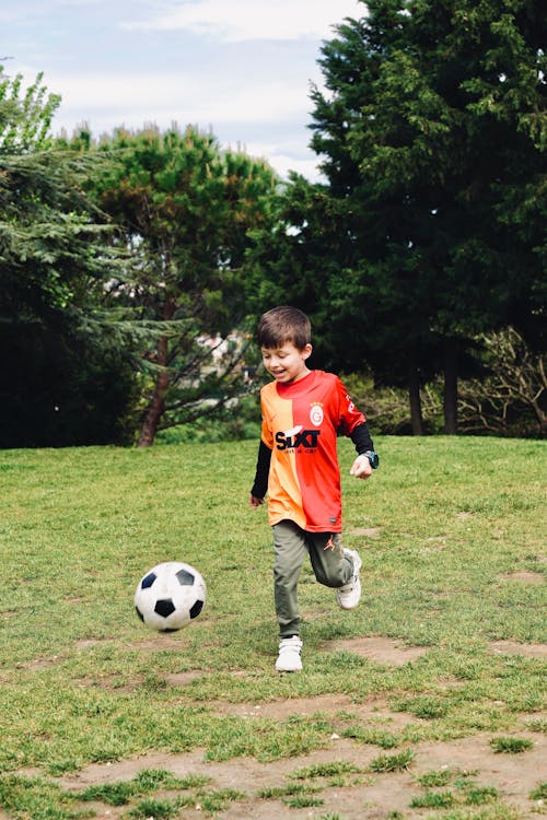 A young boy in an orange and red shirt kicking a soccer ball