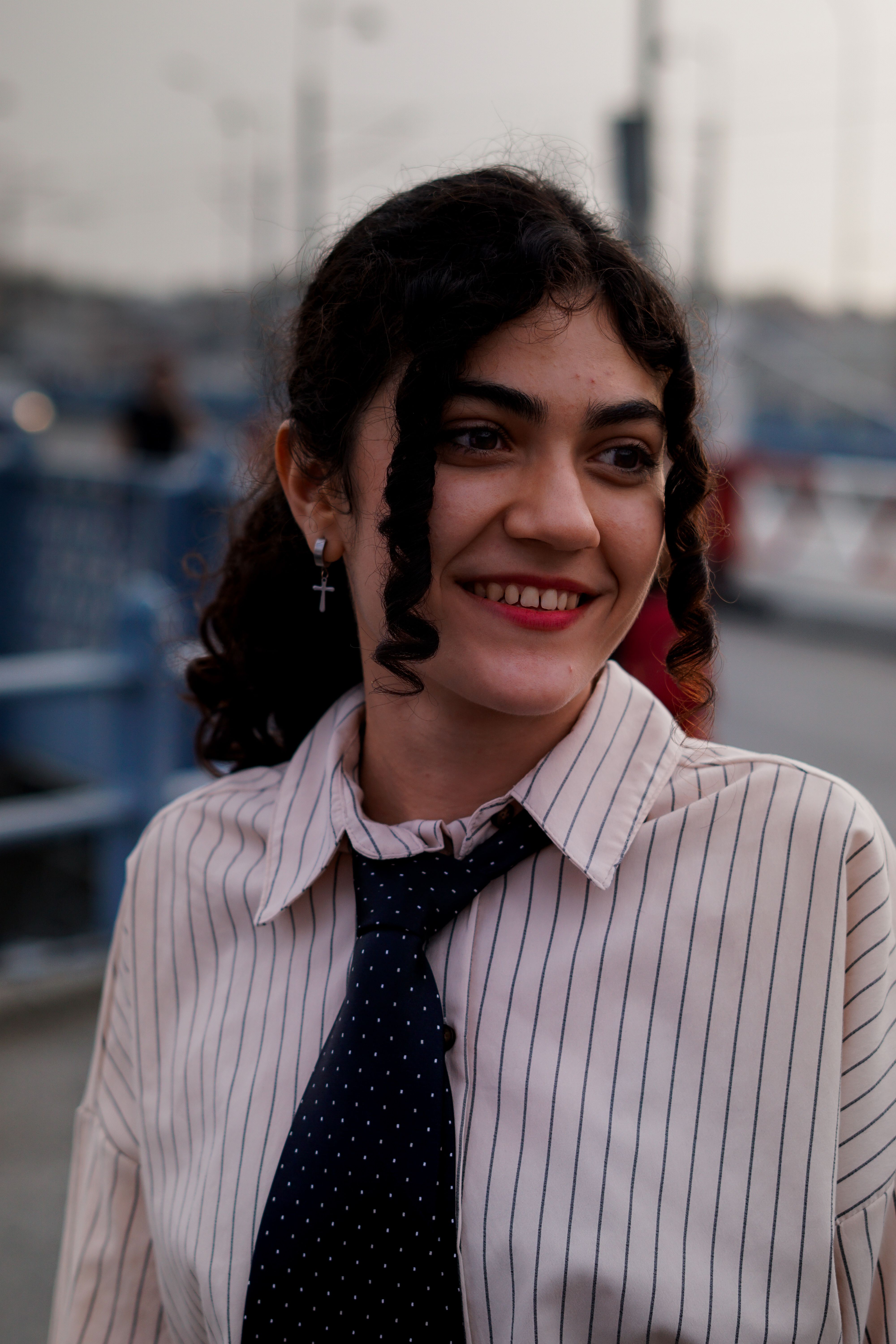 Elegant woman with curly hair and striped shirt smiling outside on a city street.