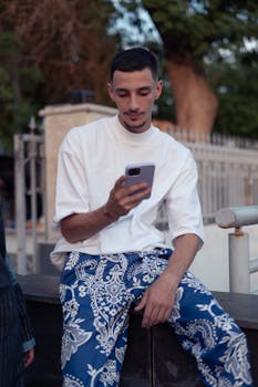 Middle Eastern man using smartphone outdoors in Baghdad, Iraq. Casual urban setting with fashionable attire.