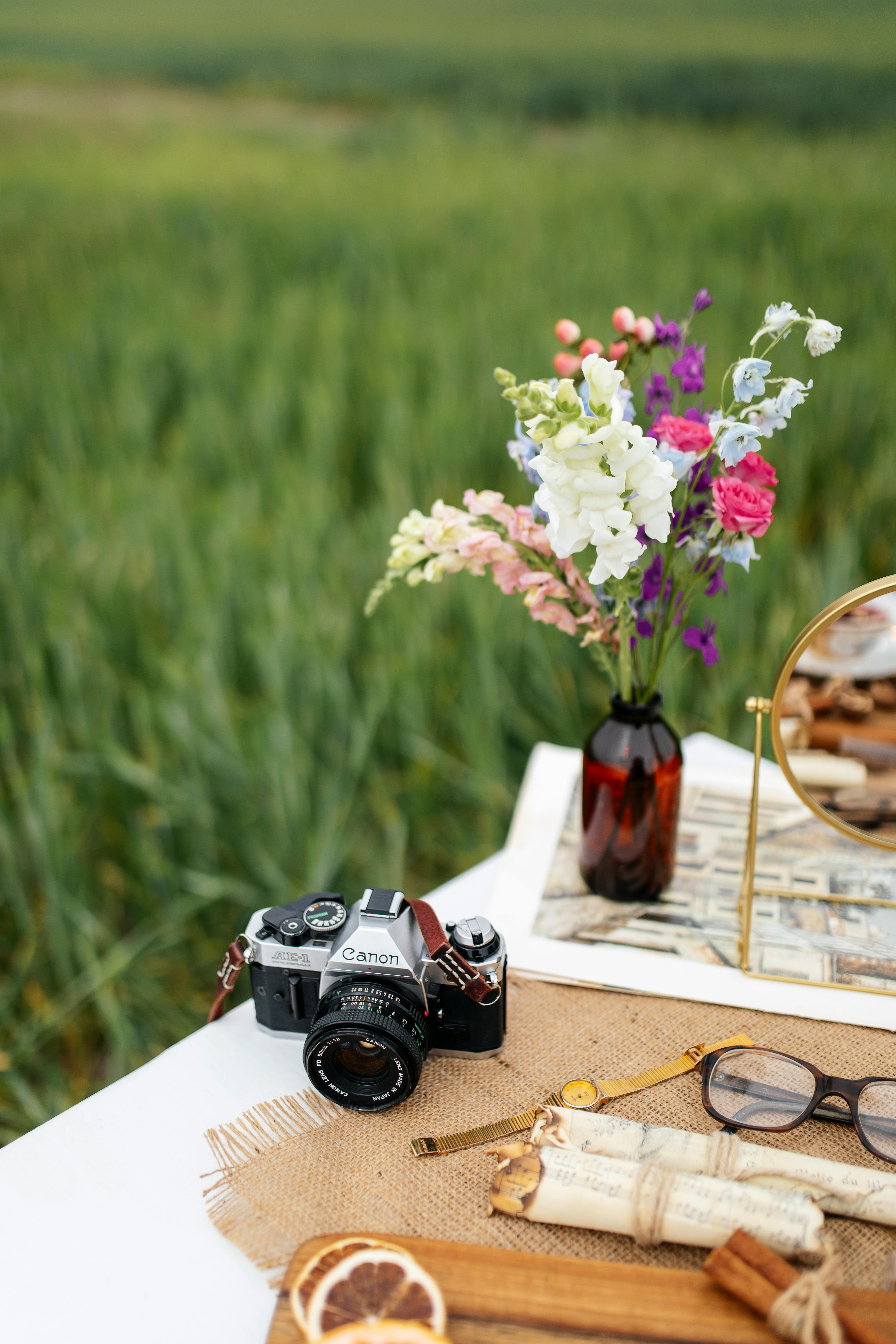 A vintage camera on a picnic table with wildflowers in a vase, set in a rural field.