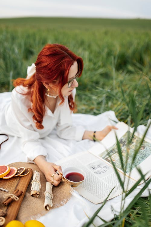 A woman with red hair reading a book in the grass