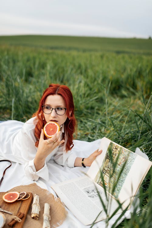 A woman with red hair and glasses is sitting in the grass with a book and a slice of fruit