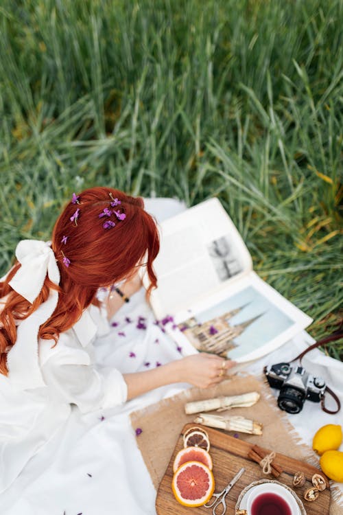 A woman with red hair sitting on the grass with a book