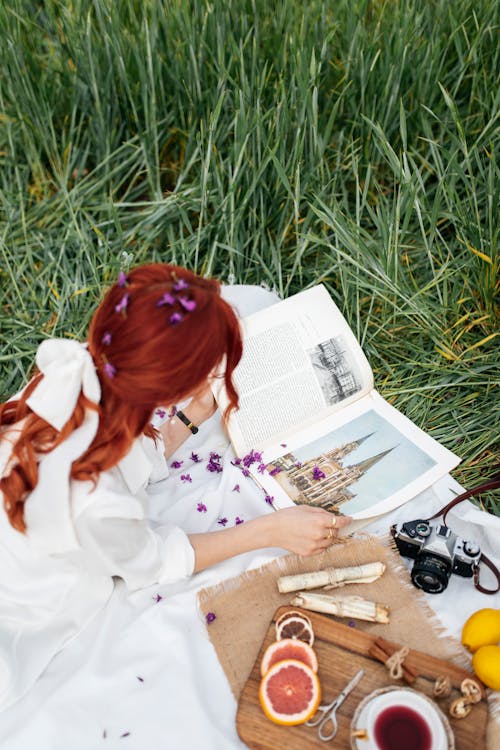 A woman with red hair sitting in the grass with a book