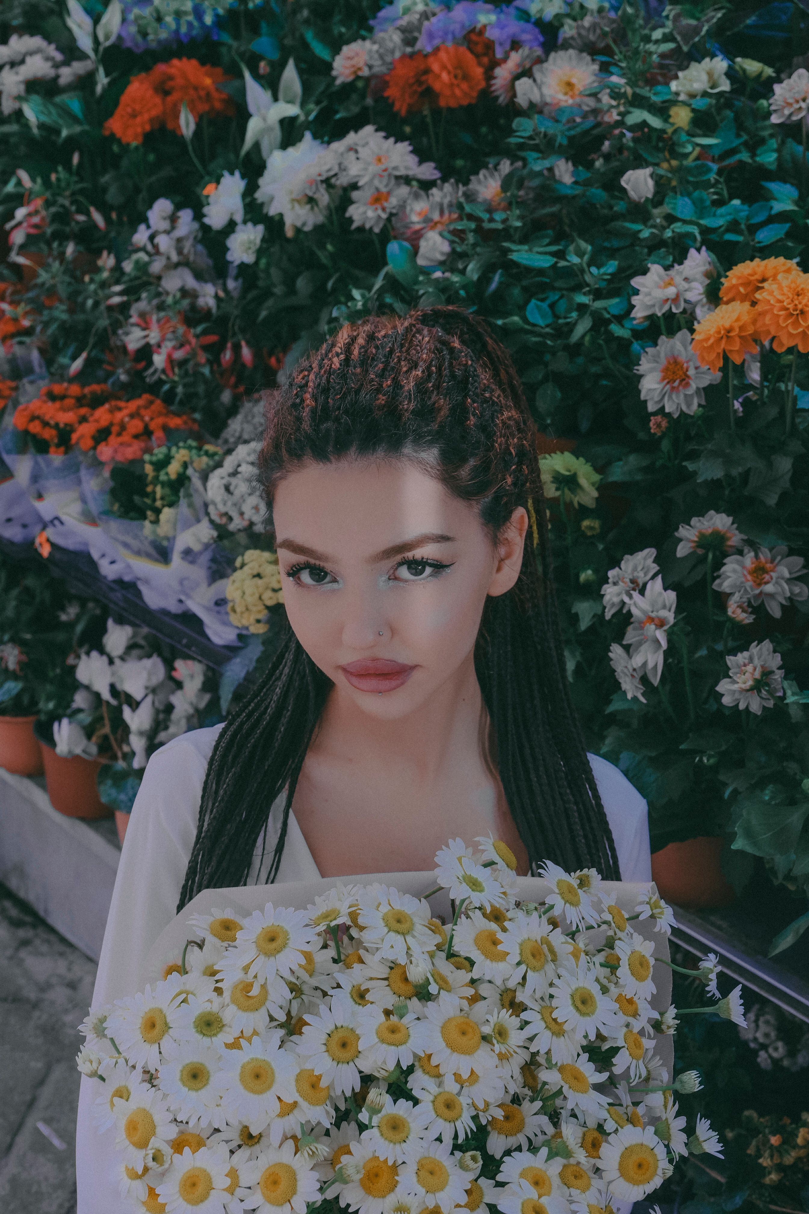 A woman with braided hair holds a daisy bouquet in front of vibrant flowers.
