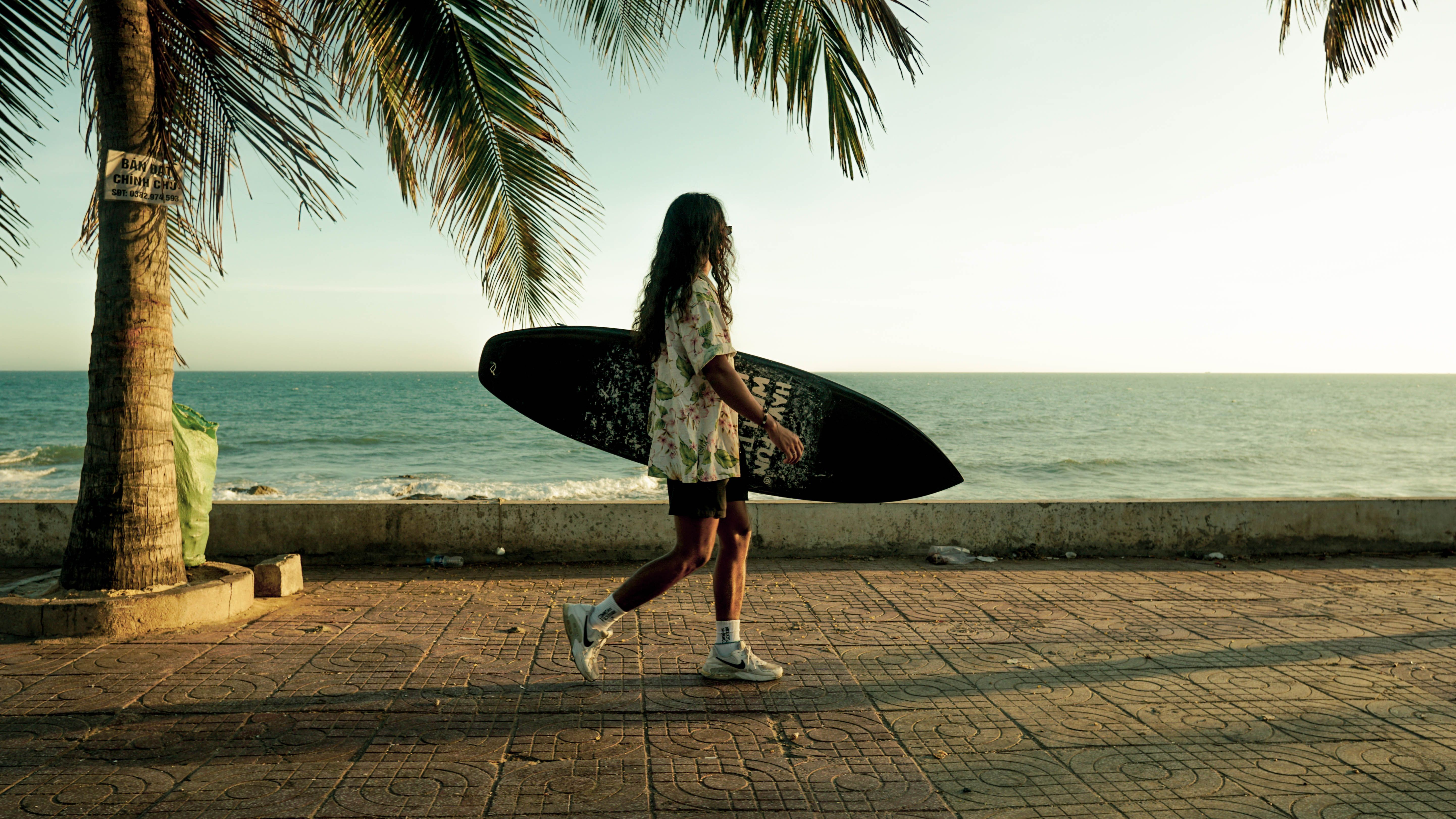 Surfer carrying a board along a palm-tree promenade by the ocean, capturing a leisure lifestyle vibe.