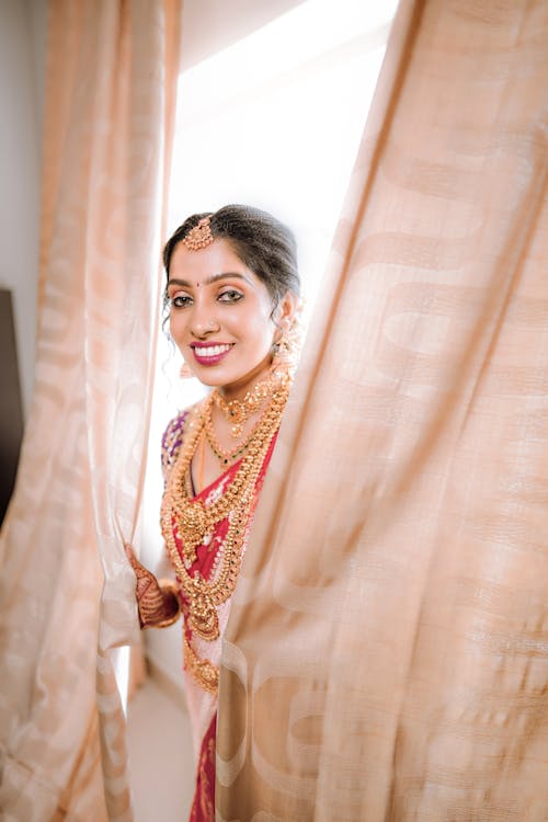 A bride in traditional indian attire smiles as she looks out of a window