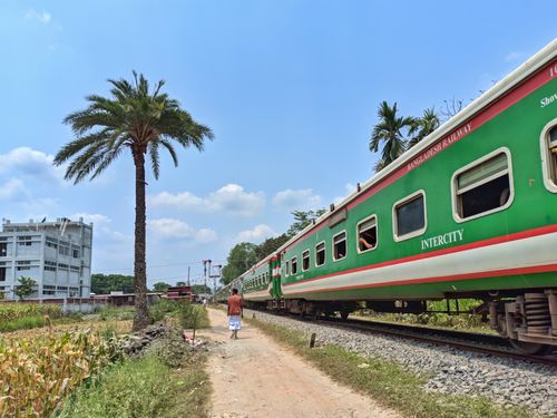 Free A man walks by a Bangladesh Railway train on a sunny day, with palm trees and buildings nearby. Stock Photo