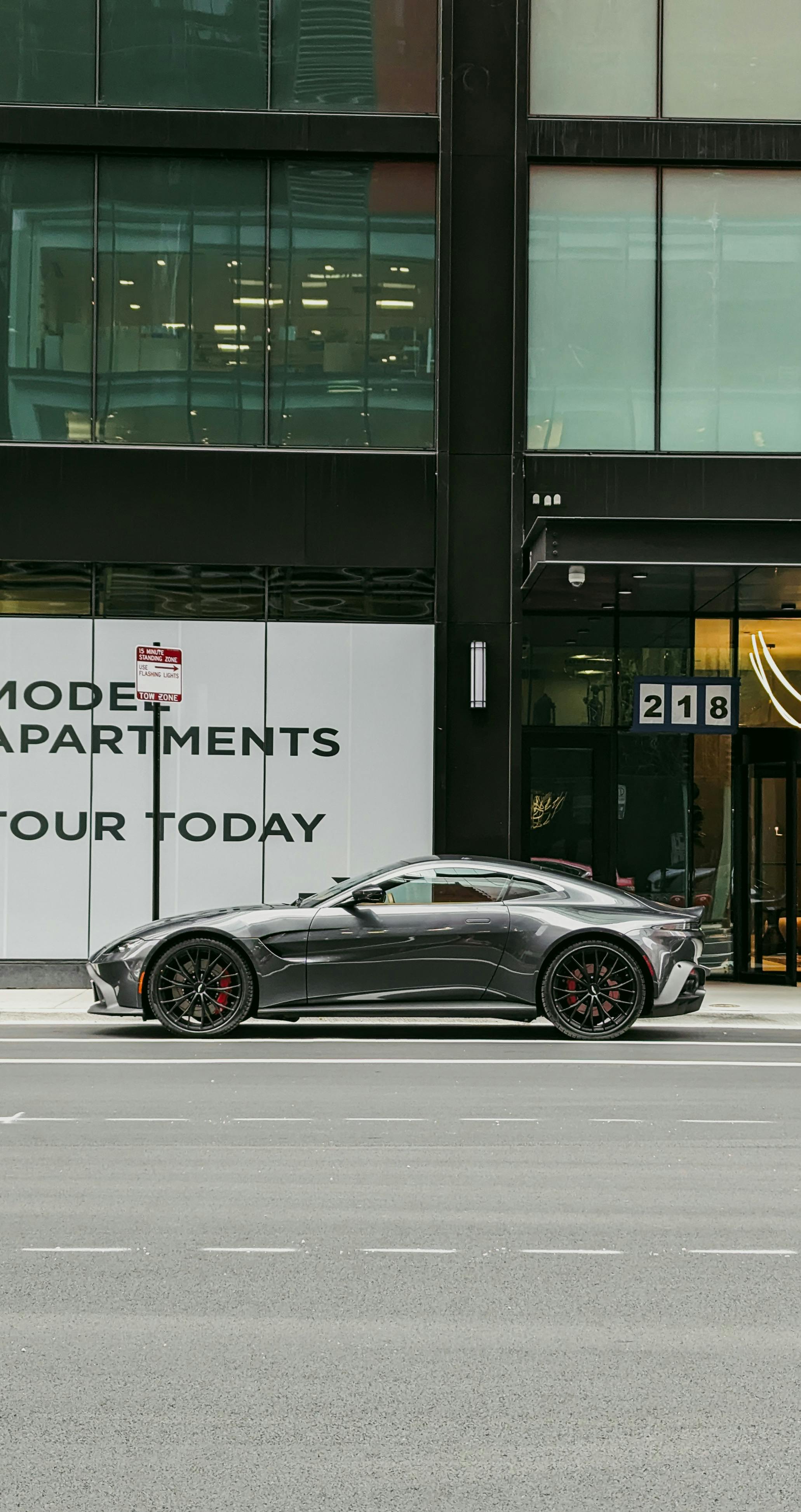 Free A sleek silver luxury sports car parked on a city street, showcasing modern architecture. Stock Photo