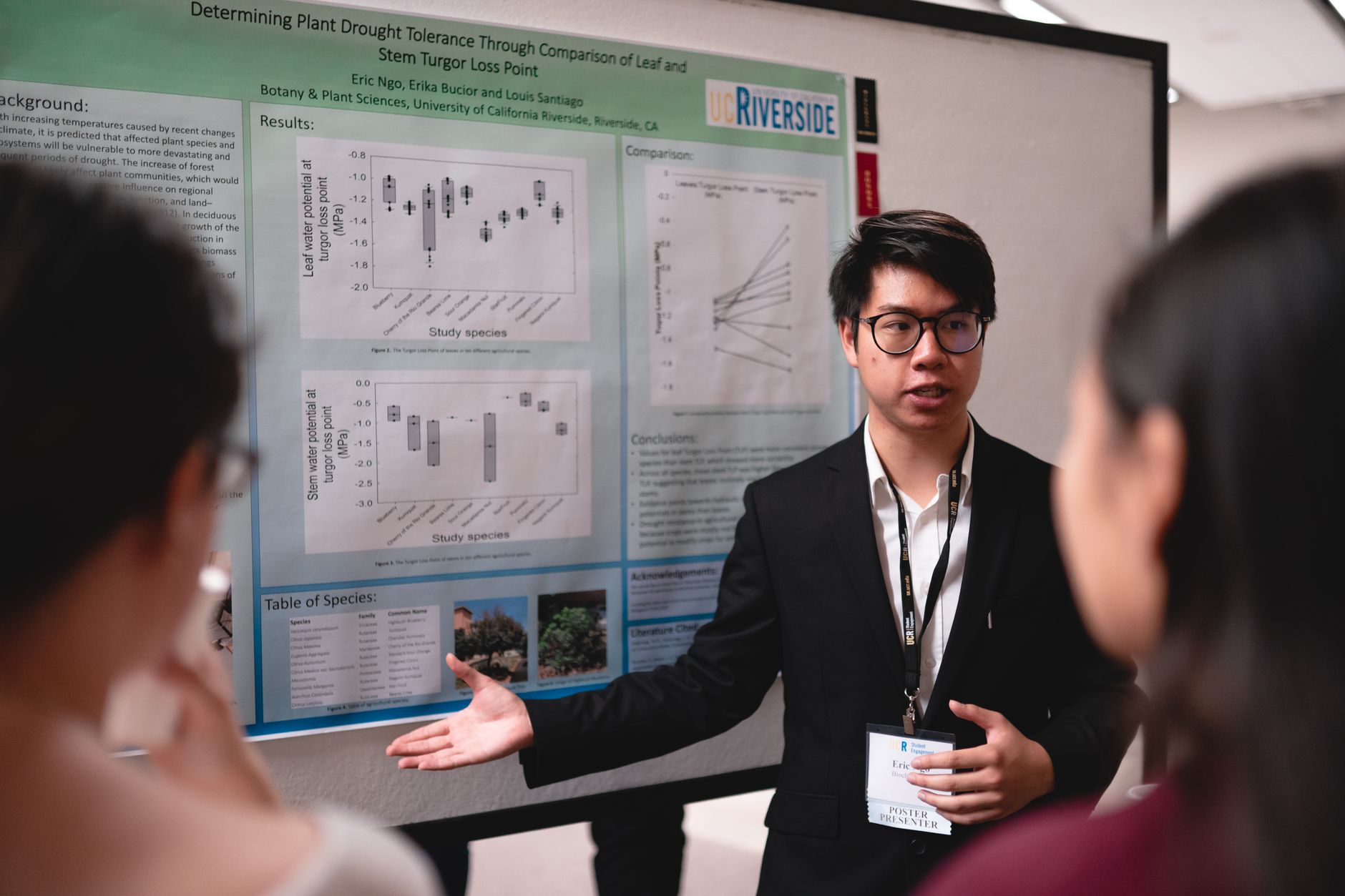 Asian man presenting scientific research on plant drought tolerance at a conference using a poster display.