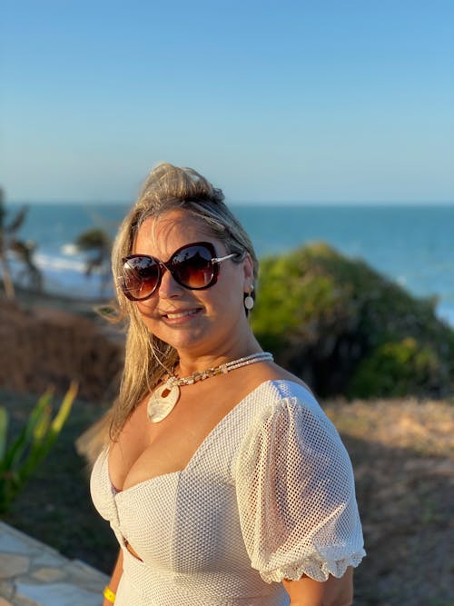 A woman in a white dress and sunglasses standing by the ocean