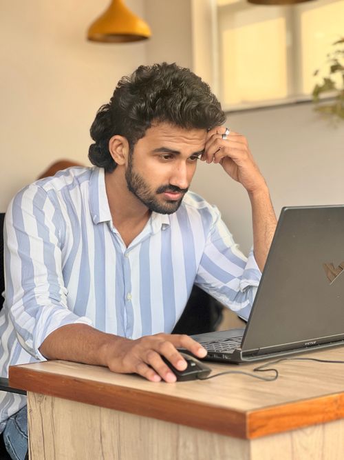 Free Young man in striped shirt focused on work using a laptop in a modern office setting. Stock Photo