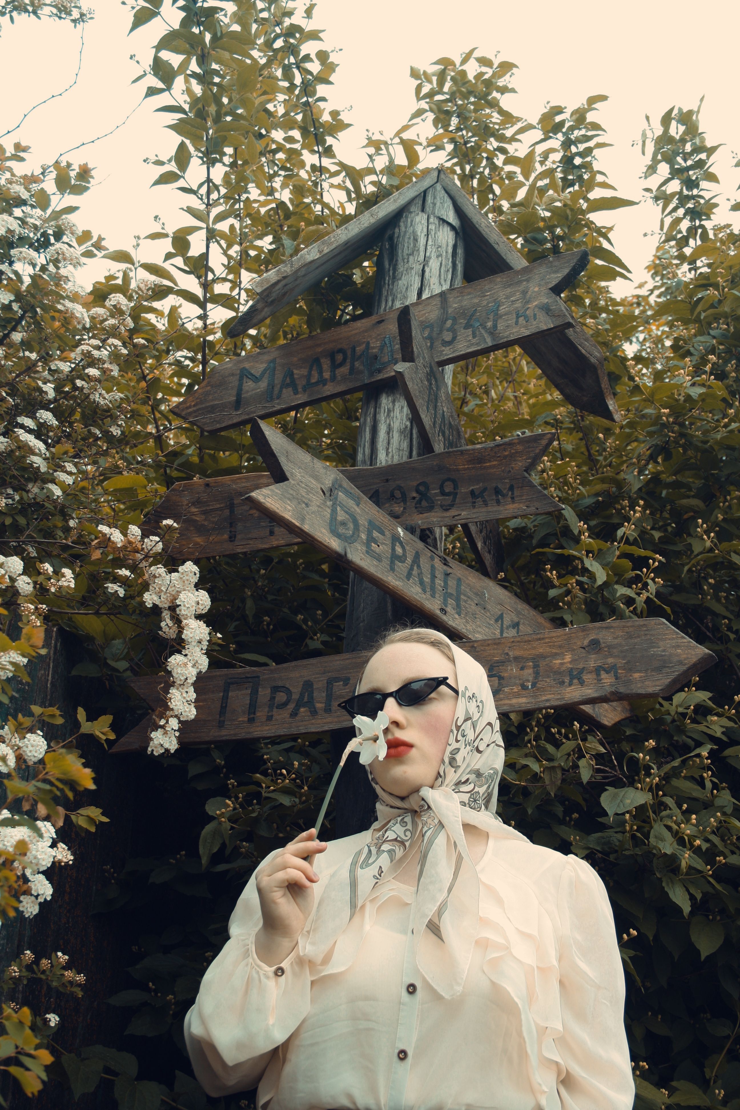 A fashionable woman with sunglasses and headscarf holding a white flower by rustic signs in a lush outdoor setting.