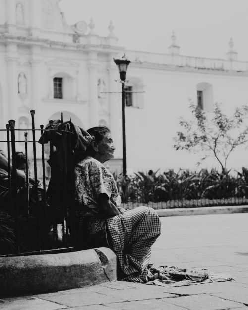 Free A contemplative scene of an elderly woman sitting by a church on a city street, captured in black and white. Stock Photo