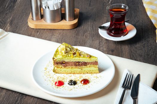 A delicious pistachio cake slice served with a glass of tea on a rustic wooden table, perfect for dessert lovers.