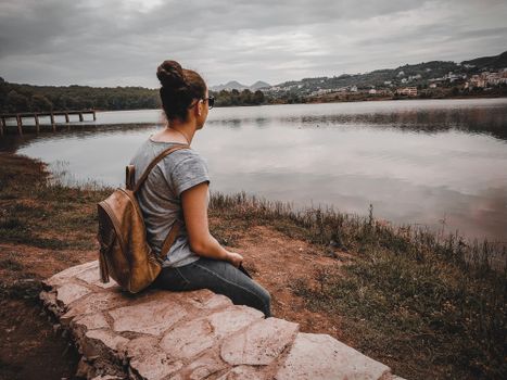 A woman sits on a stone wall by a serene lake in Albania, enjoying nature's tranquility.