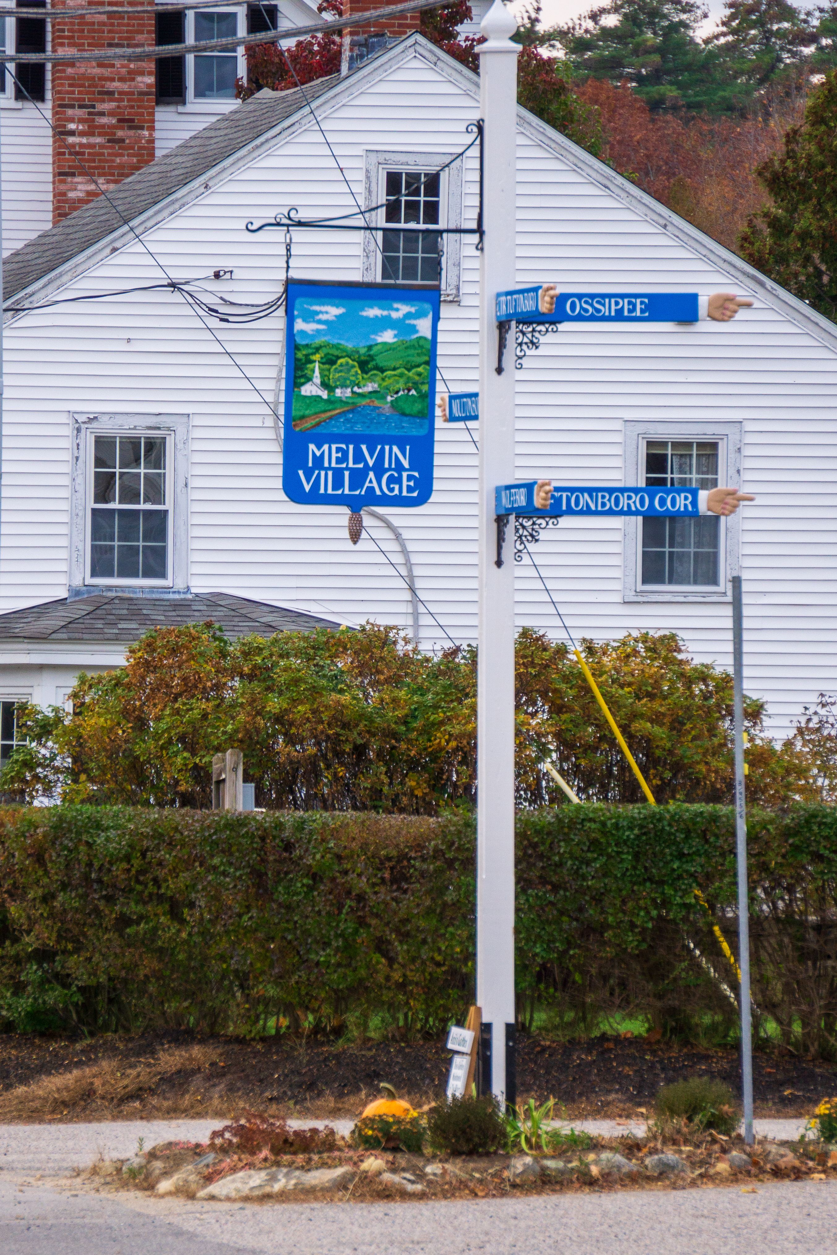 Charming Melvin Village signpost with autumn foliage and historic white home in background.