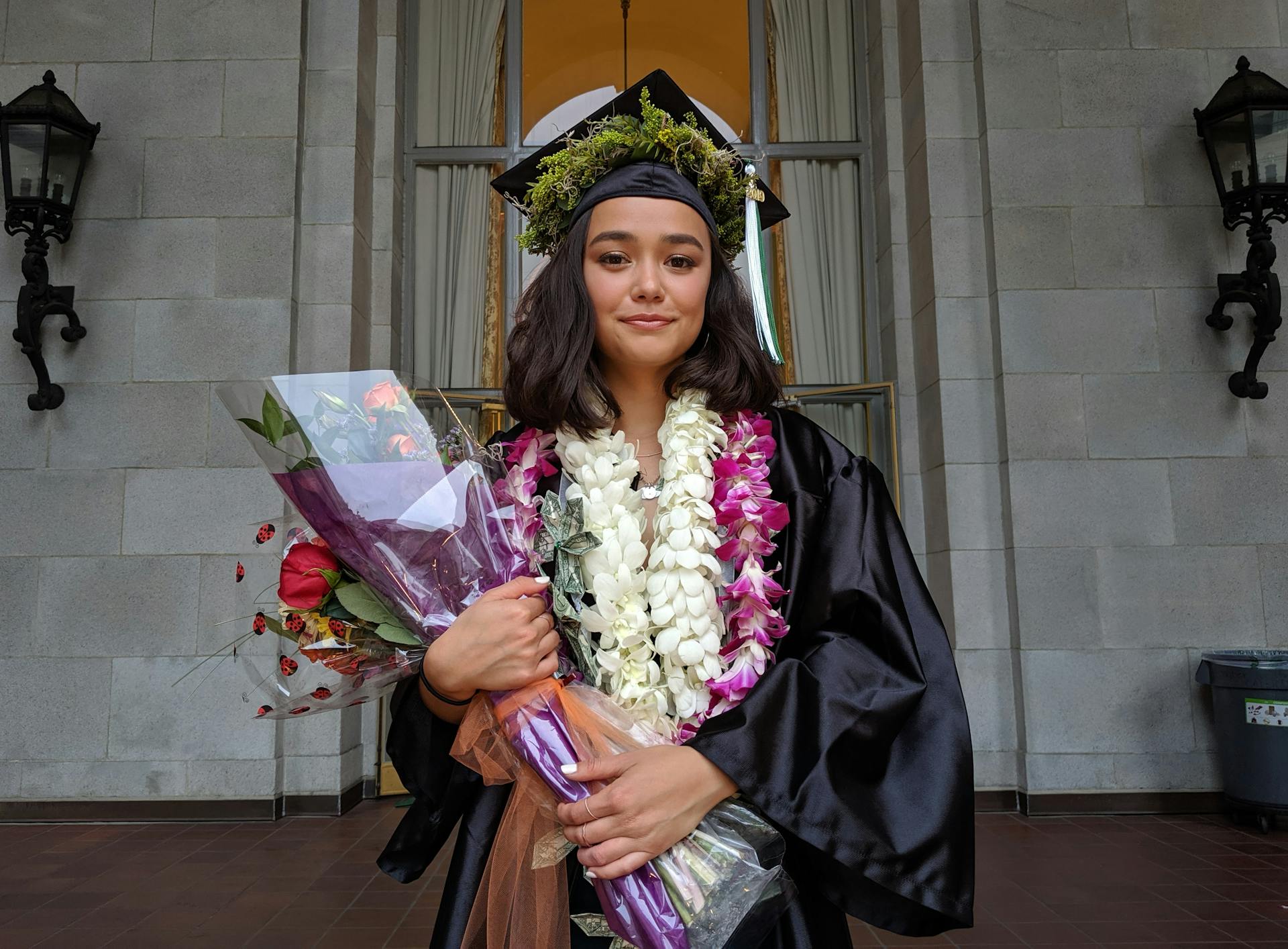 A proud graduate wearing a cap and gown, holding a lei and flower bouquet, symbolizing achievement.