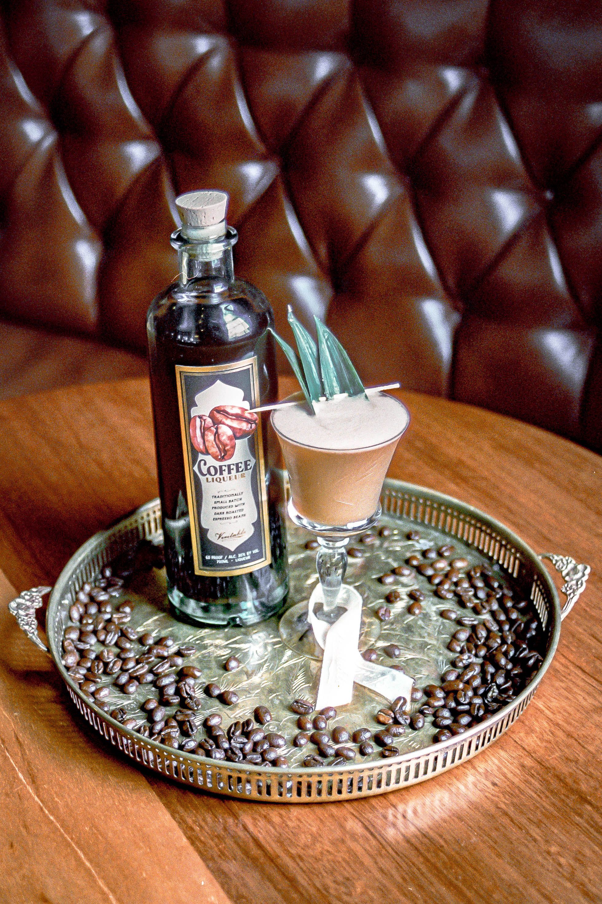 A dark bottle of coffee liqueur poured over ice in a rocks glass, surrounded by whole coffee beans on a wooden counter.