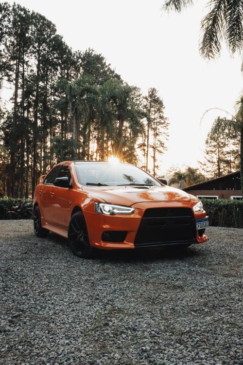 Free Striking orange Mitsubishi Lancer parked outdoors against a scenic sunset backdrop. Stock Photo