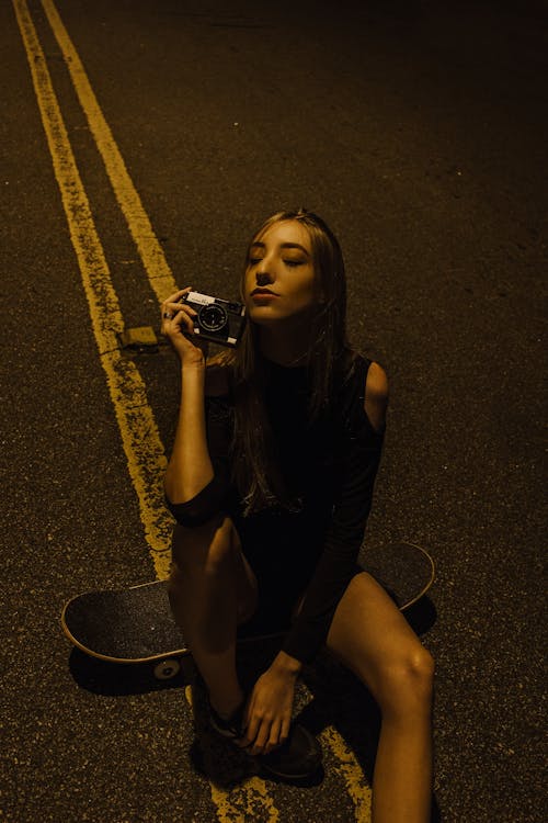 그녀의 눈을 감고 카메라를 들고있는 동안 도로 한가운데 스케이트 보드에 앉아있는 여자의 사진
