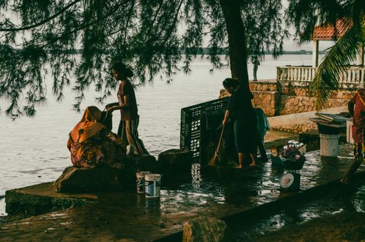 Women tending to chores by a serene waterfront with trees and distant scenery.