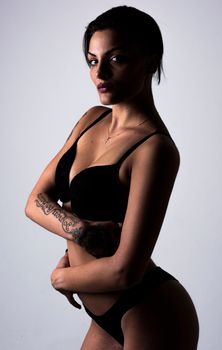 Studio portrait of a woman wearing a black bikini with tattoos on her arm against a gray background.