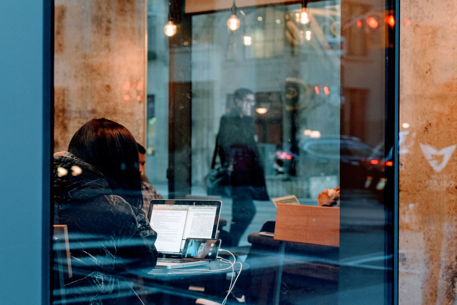 Seen from the street through a large café window, a person with dark hair sits at a counter working on an open laptop, a phone charging beside it, while the glass reflects a man in a dark coat walking past on the pavement outside