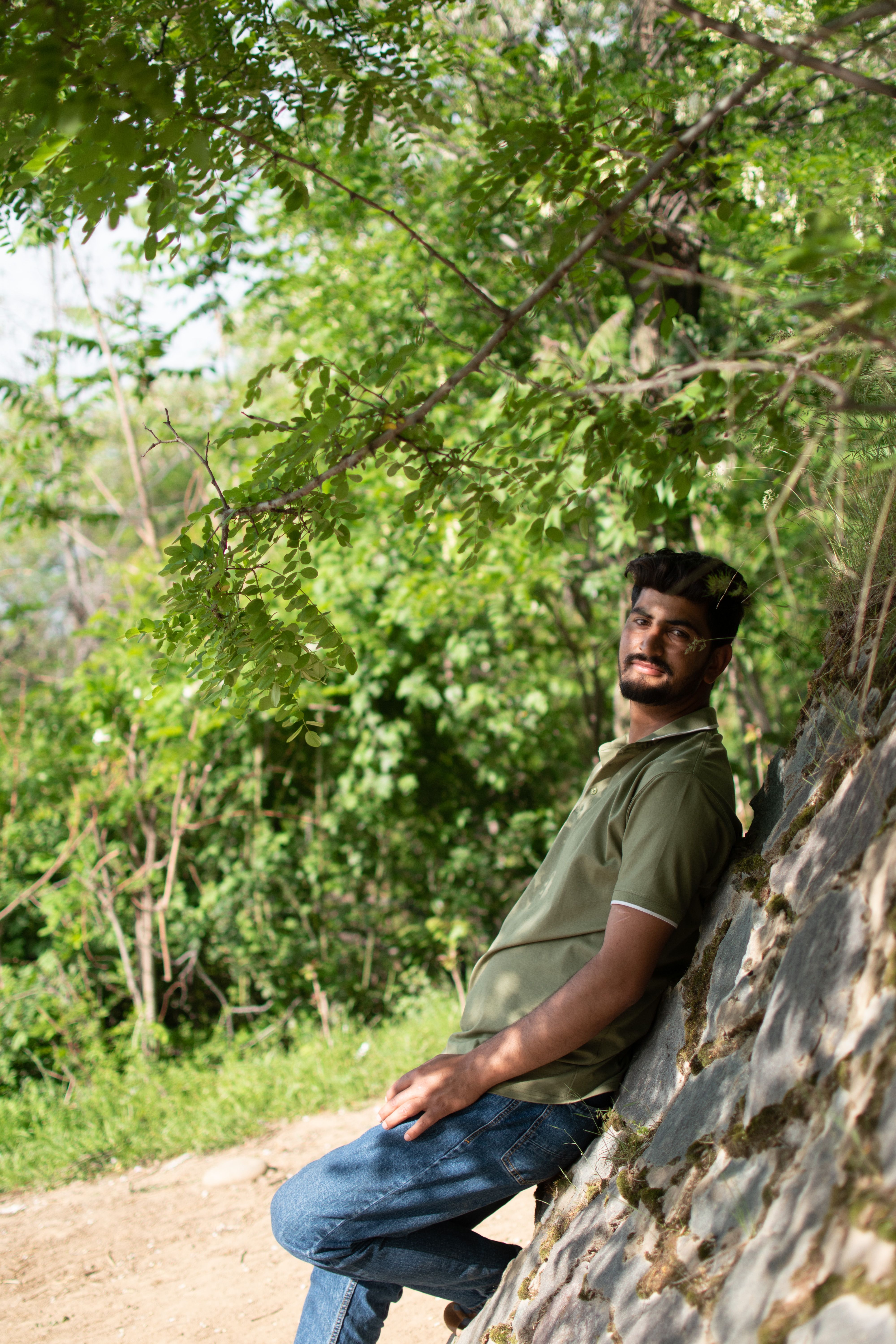 A man in a green t-shirt leans against a stone wall in a lush outdoor setting.
