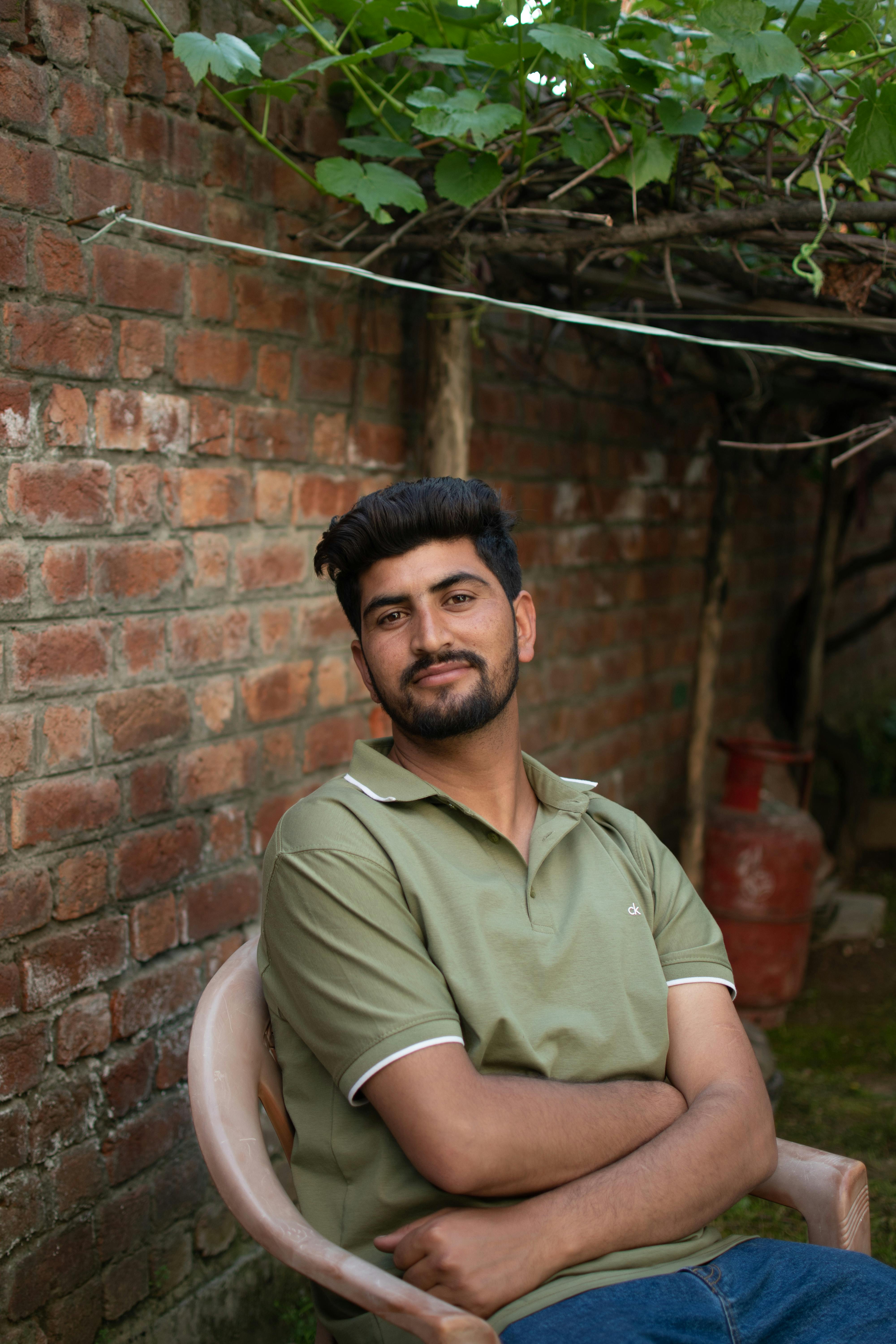 A young South Asian man sits relaxed outdoors against a brick wall with greenery overhead.