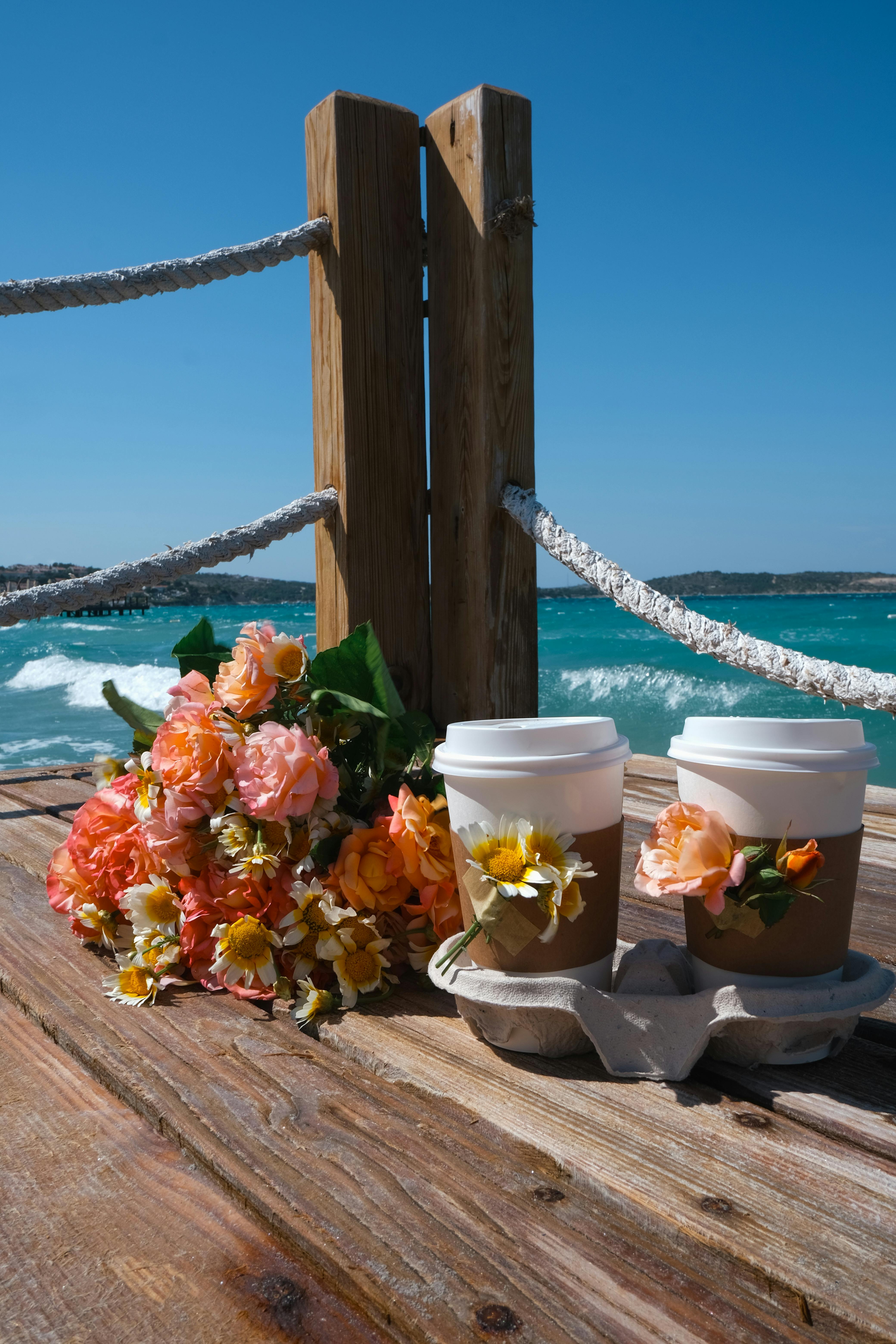 Two coffee cups and a flower bouquet on a pier with a sea view, perfect for relaxation.