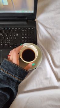 A person with turquoise nails holds a coffee cup near a laptop on a white bedspread.