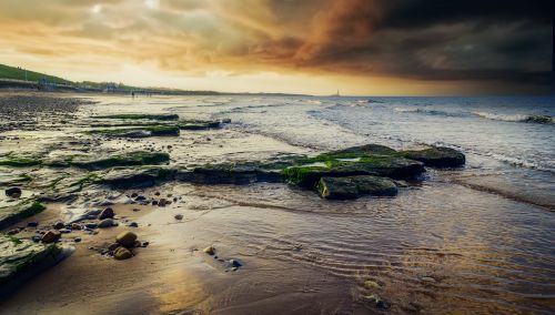 Free Serene beach with dark clouds, lighthouse, and vivid sunset overlooking the ocean waves. Stock Photo
