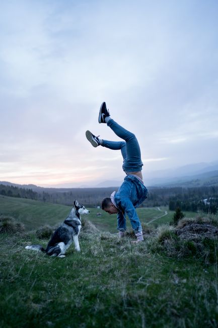 Man performs handstand beside husky dog on grassy hill at sunset, embracing freedom.