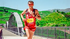 在一个阳光明媚的日子里，一位身穿红色衣服的男性马拉松运动员站在一座风景优美的桥上