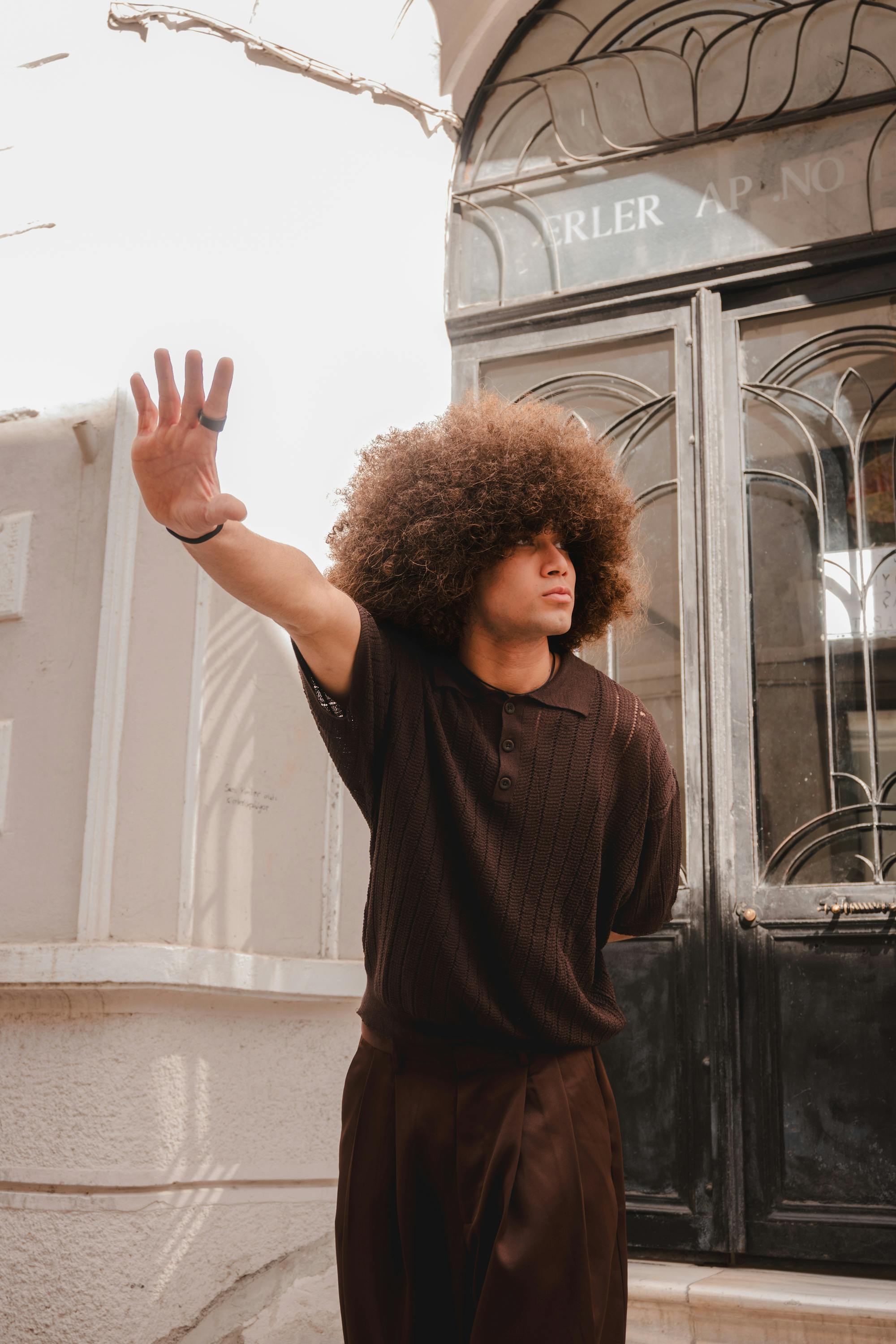 Fashionable young man with curly hair posing outdoors in a brown outfit, hand raised against sunlight.