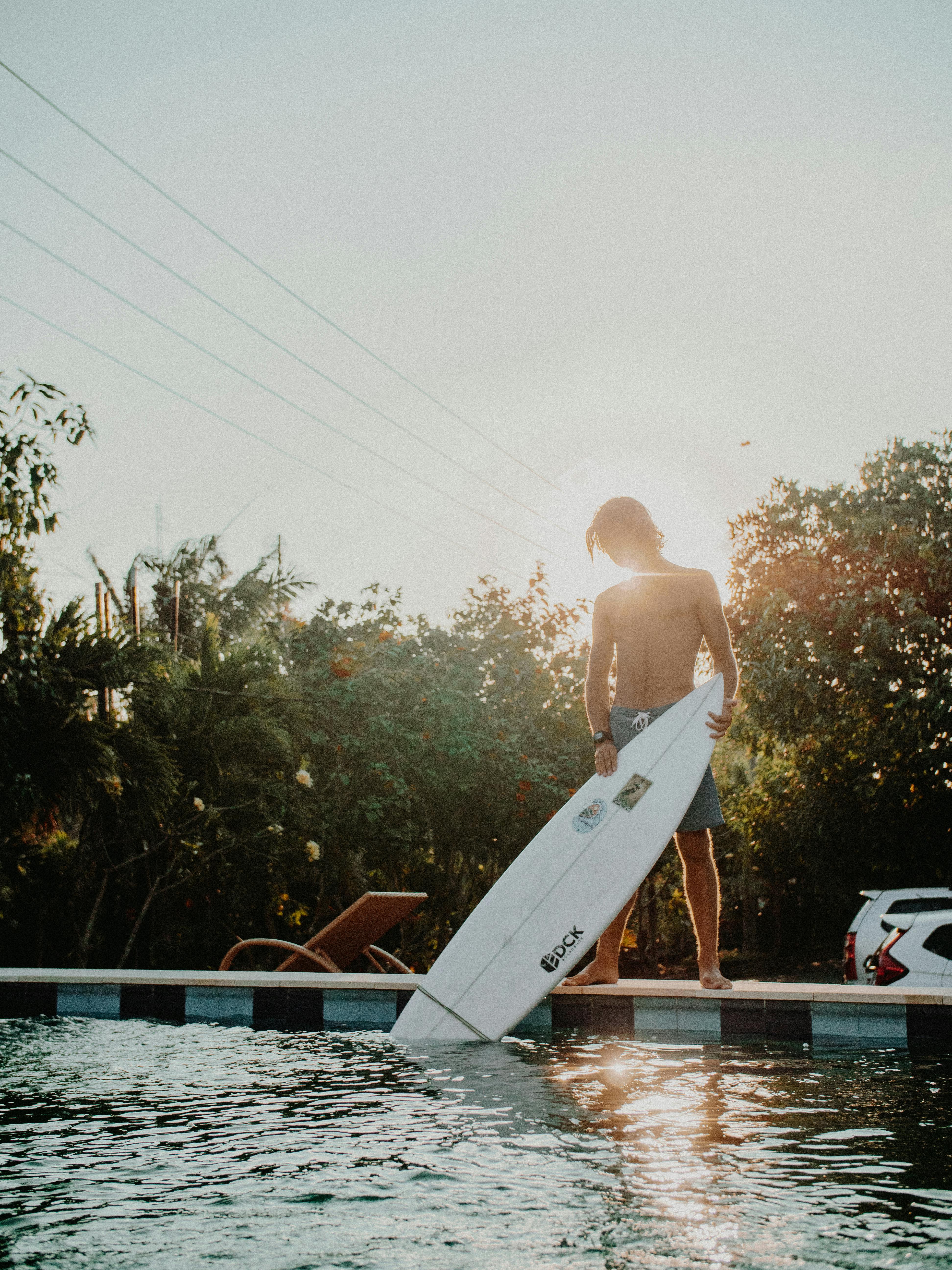 一名年轻人拿着冲浪板，站在阳光照射的户外泳池边
