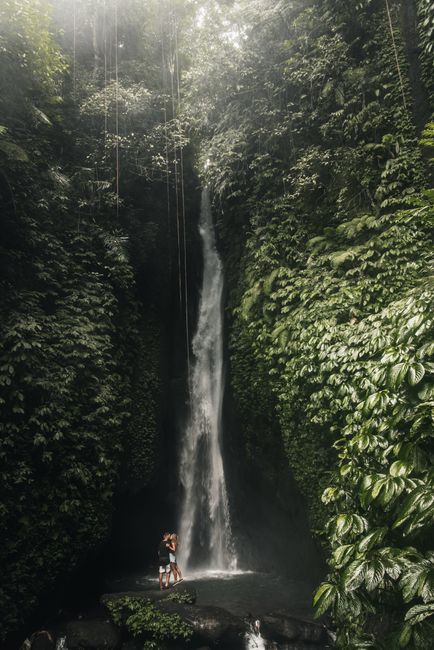 A couple embraces at the base of a lush tropical waterfall surrounded by dense greenery.