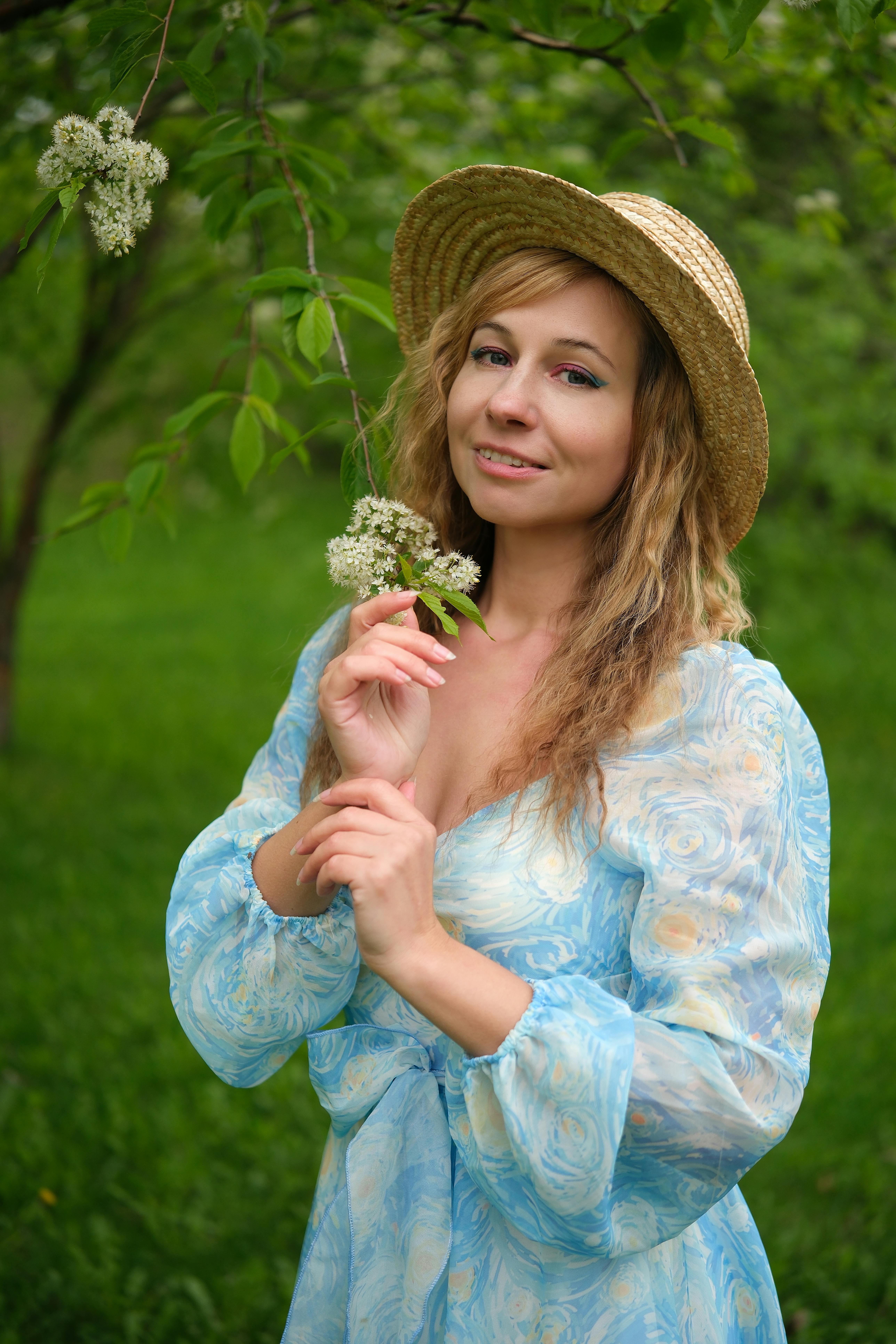 Beautiful woman in floral dress and straw hat posing with white flowers outdoors.