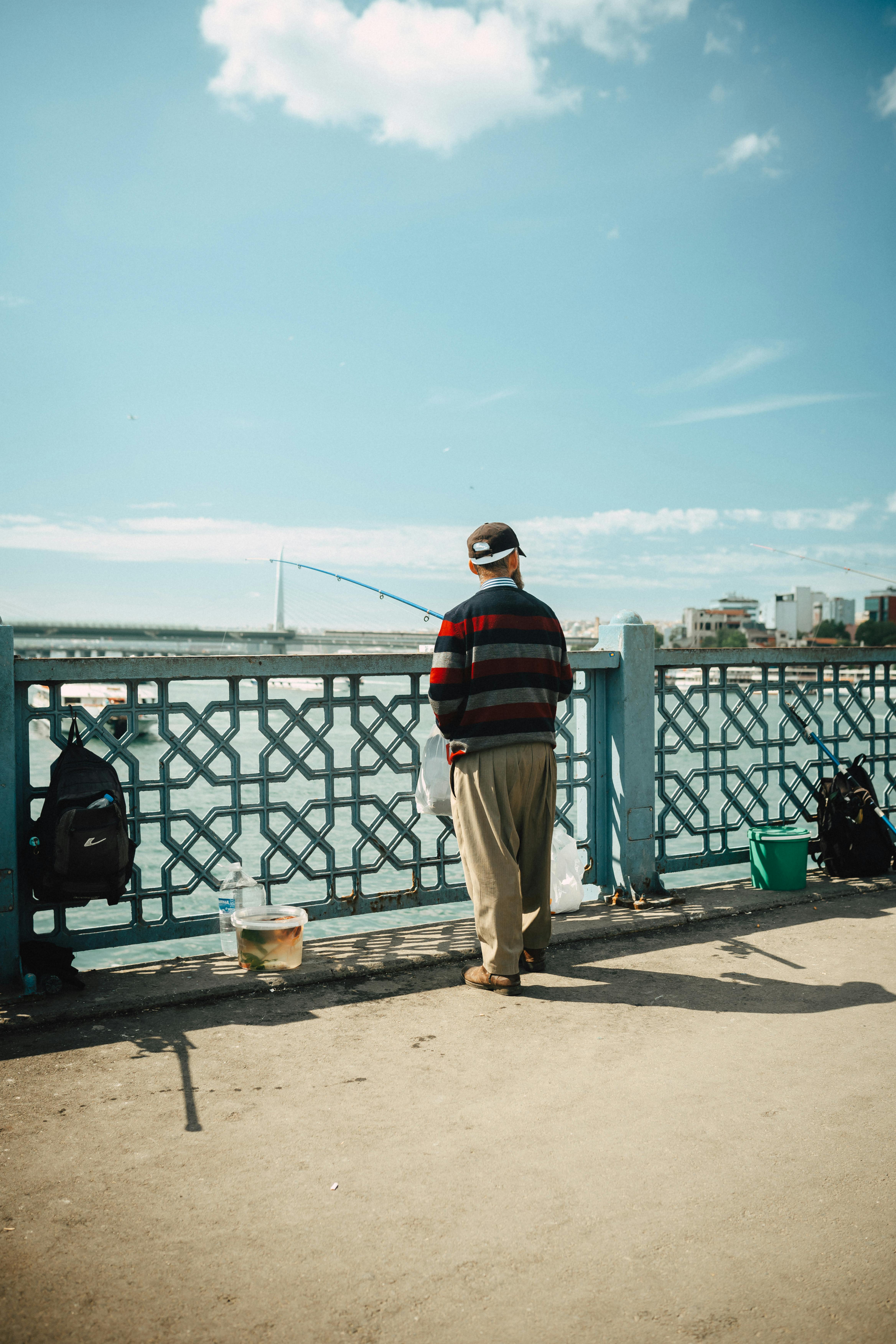 A man fishing on an urban bridge with bags and fishing gear under a clear blue sky.