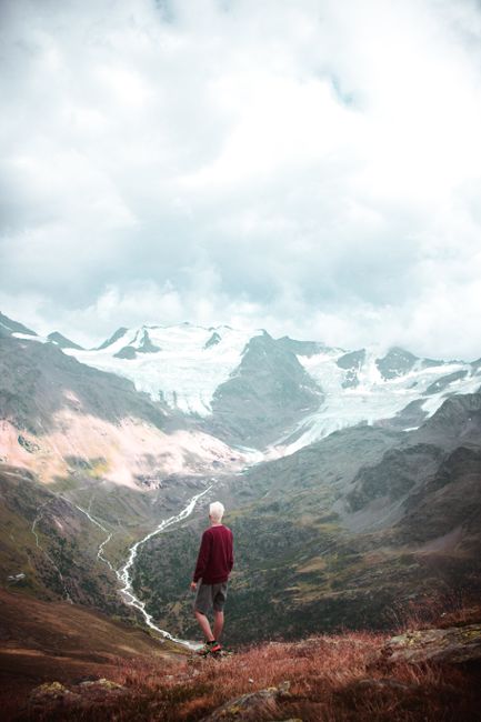 A lone hiker in shorts gazes at a stunning alpine mountain vista with glaciers under a cloudy sky.
