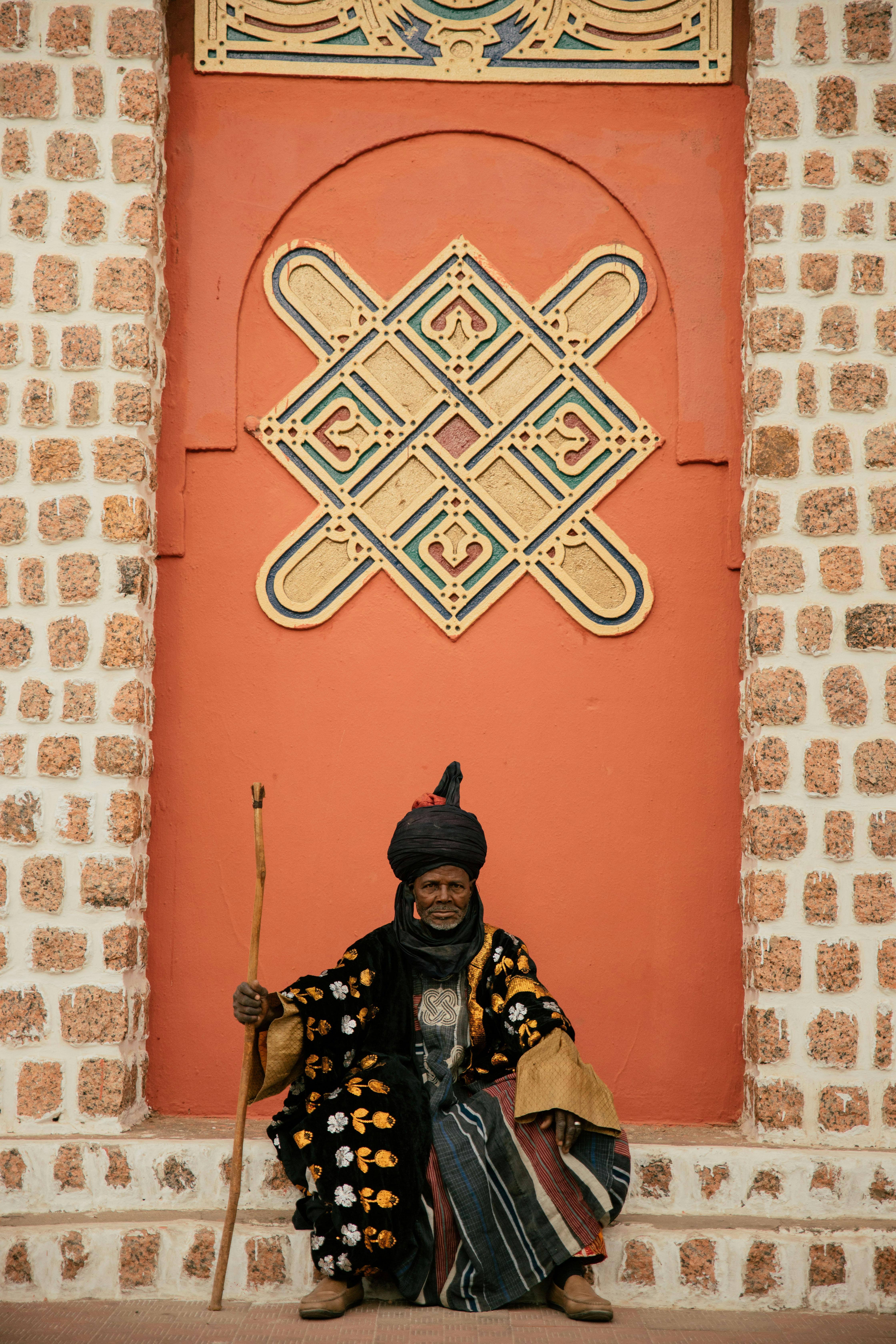 An adult dressed in cultural attire sitting against ornate Nigerian architecture, celebrating tradition.