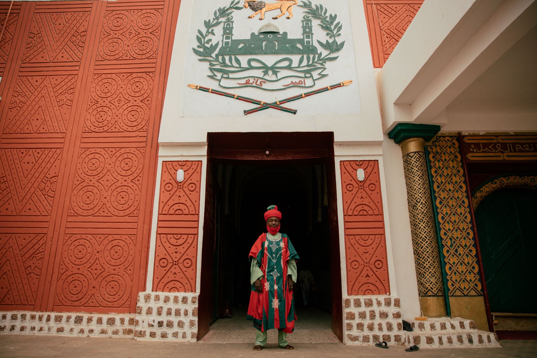 A man in traditional attire stands at a historic Nigerian landmark entrance.