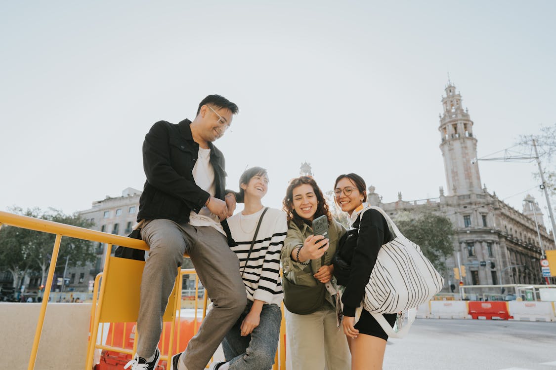 Group of four young friends taking a selfie while sitting on yellow railings with a historical tower building in background.