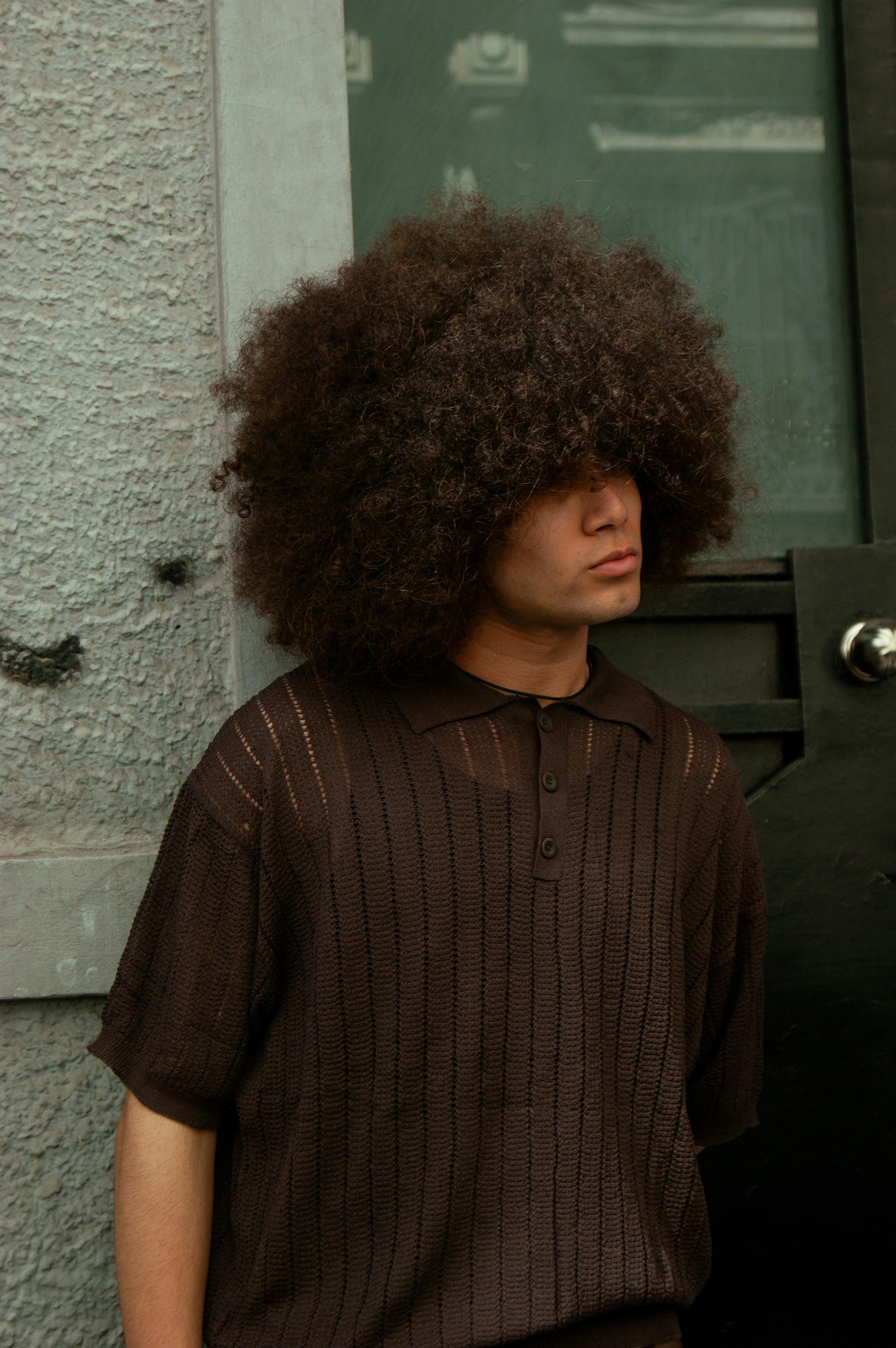 Young man with a voluminous afro hairstyle stands casually against textured wall.