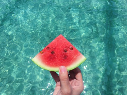 Free Hand Holding a Slice of Watermelon With Blue Swimming Pool Water in the Background Stock Photo
