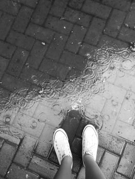 Black and white image of feet in sneakers standing on wet paving stones with rain reflections.