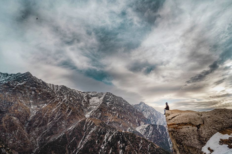 A lone adventurer sits atop a Himalayan peak with dramatic clouds overhead, creating a stunning winter landscape.
